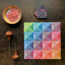 Load image into Gallery viewer, A square print on paper consisting of a geometric deign with triangles, squares and stripes in an ombre of rainbow colors. also shown are two carving tools, the stamp which was used to make the print and a dish of the carving scraps.