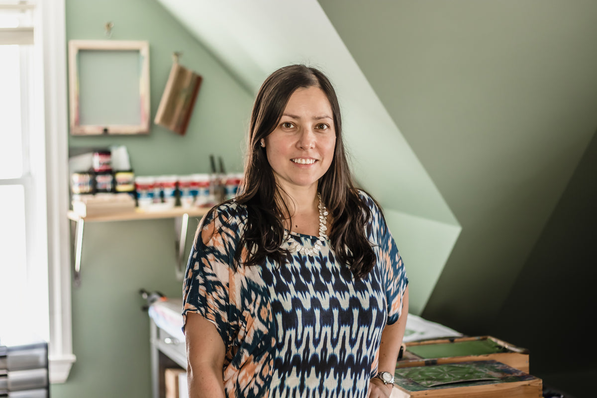 Photo of Susana McDonnell standing in her printmaking studio with printing table and inks behind her.