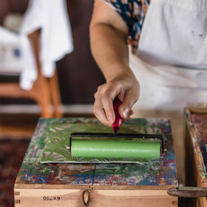 Susana McDonnell's hand holding a brayer with light green ink and rolling the ink onto a piece of linoleum with a geometric design carved into it.