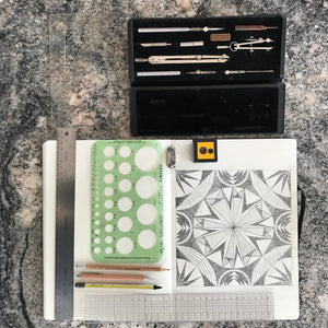 A photograph of the tools that Susana McDonnell of LinoCave uses to create her designs. There is a vintage compass set, a green circle tracer, rulers, a variety of different pencils, a pencil sharpener, an eraser and a sketchbook with a geometric design.