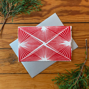 A hand block printed 4"x6" card with red ink on white paper. Features a fine-line design. An A4 metallic silver envelope is placed behind it. It is surrounded by greenery on a distressed wood background.