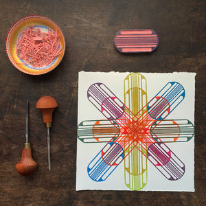 Am 8"x8" print on paper with a star-shaped hand block printed pattern of an oval and striped design. Printed in a vivid color scheme. also shown are the oval shaped block, dish of scraps and carving tools.
