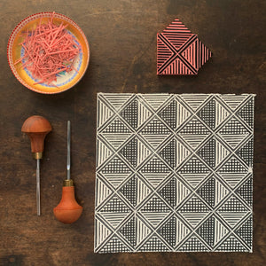 A square print on paper consisting of a geometric deign with triangles, squares and stripes in black and white. also shown are two carving tools, the stamp which was used to make the print and a dish of the carving scraps.