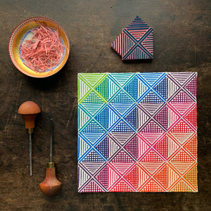A square print on paper consisting of a geometric deign with triangles, squares and stripes in an ombre of rainbow colors. also shown are two carving tools, the stamp which was used to make the print and a dish of the carving scraps.