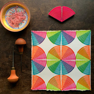 an image of an 8"x8" art print in a circular pattern with line elements and vivid color scheme the fan-shaped block, dish of carving scraps and carving tools are also pictured.